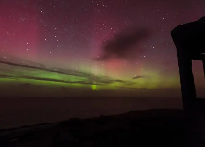 Aurora borealis illuminates the night sky with green and pink hues over a coastal landscape with a silhouette of a structure on the right.