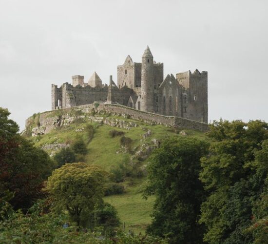 An ancient, medieval castle with multiple towers, perched atop a grassy hill in Ireland, surrounded by green foliage under a cloudy sky.