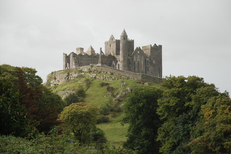 An ancient, medieval castle with multiple towers, perched atop a grassy hill in Ireland, surrounded by green foliage under a cloudy sky.