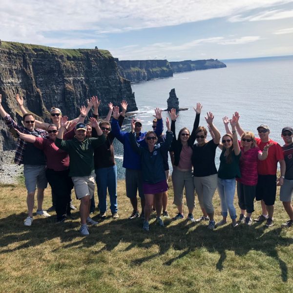 Group of people posing with raised hands in front of the cliffs of moher on a clear day.