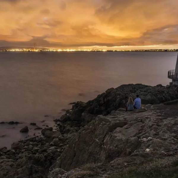 Two individuals sitting on a rocky outcrop by the water, overlooking a cityscape at dusk.