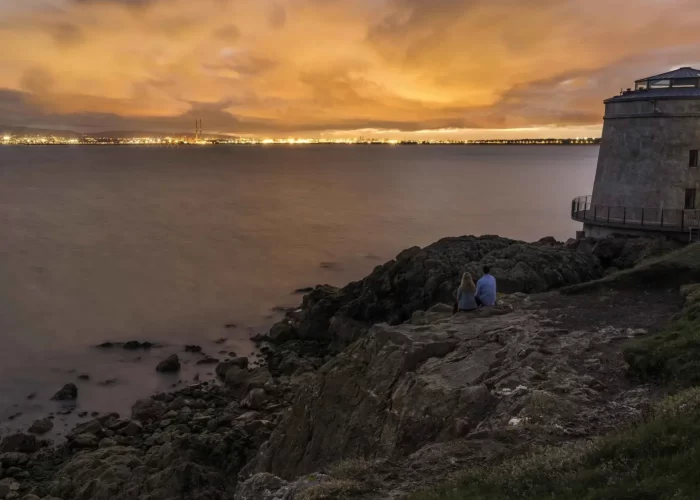 Two individuals sitting on a rocky outcrop by the water, overlooking a cityscape at dusk.