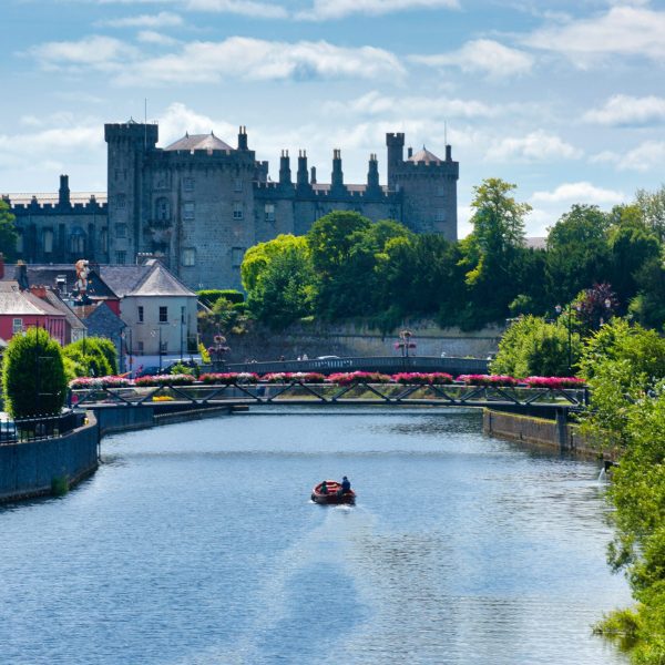 Historic castle overlooking a river with a small boat and blooming flowers along the banks.