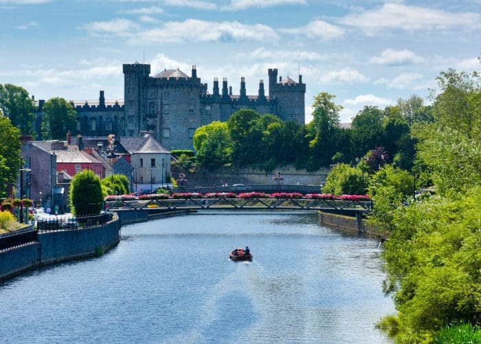 Historic castle overlooking a river with a small boat and blooming flowers along the banks.