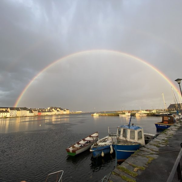 A vivid double rainbow arching over a calm harbor with boats and waterfront buildings under a stormy sky.