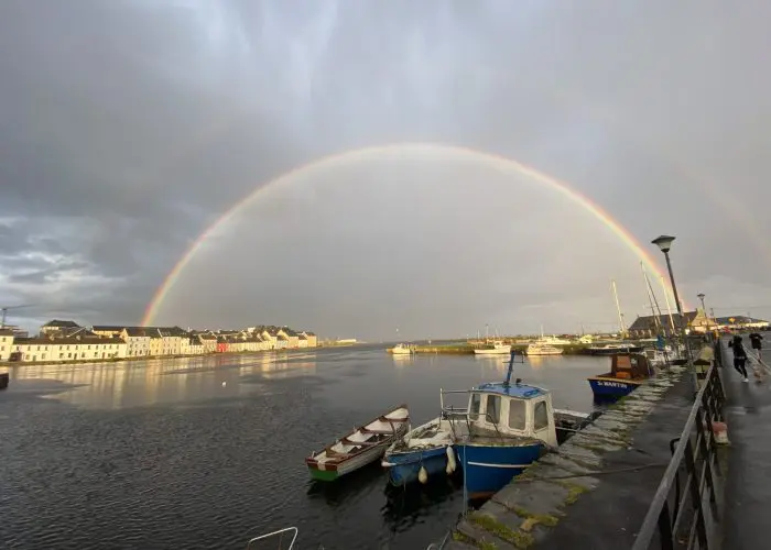 A vivid double rainbow arching over a calm harbor with boats and waterfront buildings under a stormy sky.
