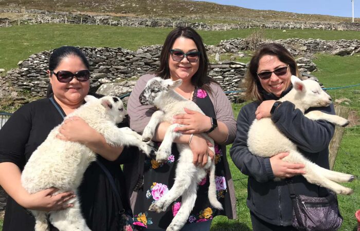 Three women smiling and holding lambs in a rural setting in Ireland.