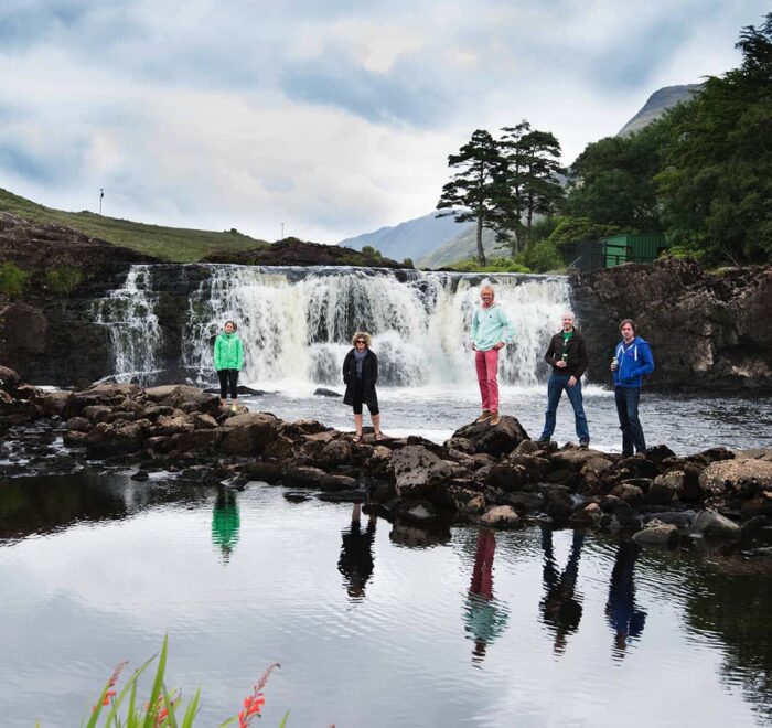 A group of people on a private tour walking across stepping stones in front of a waterfall with lush greenery in Ireland's background.
