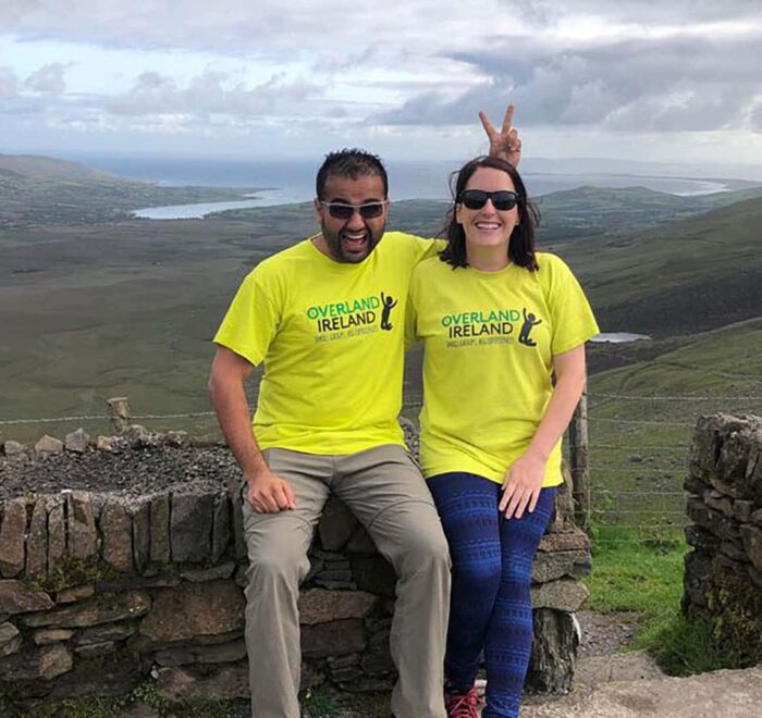 Two people wearing yellow "Ireland Tours" t-shirts smiling and posing for a photo with a scenic landscape and water body in the background. One person is playfully making a "bunny ears