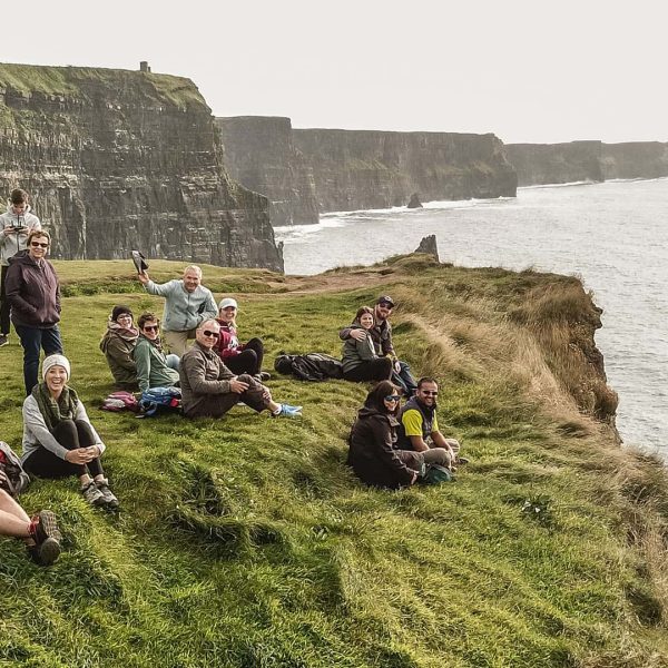 Group of people sitting on grassy cliff edge with a view of coastal cliffs in the background.