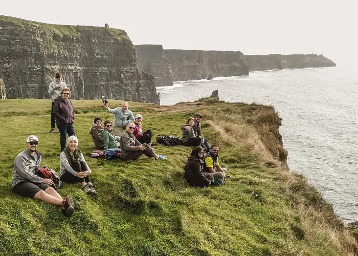 Group of people sitting on grassy cliff edge with a view of coastal cliffs in the background.