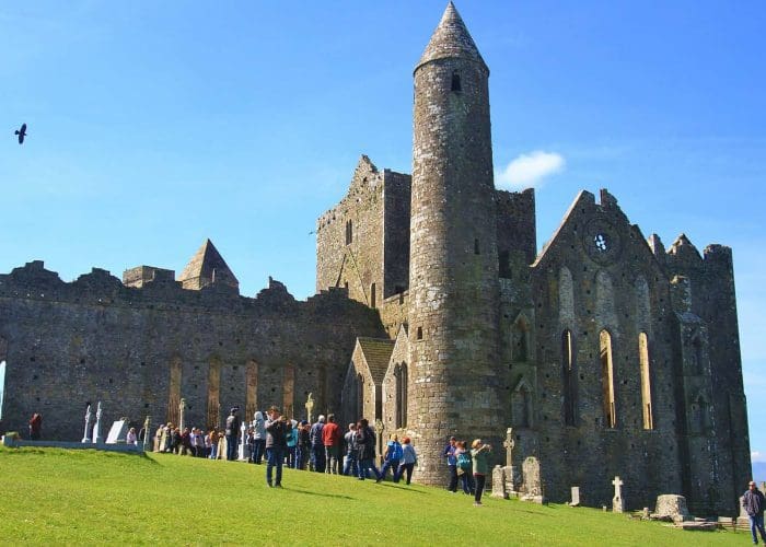 Tourists visiting the ruins of a medieval cathedral with a round tower on a sunny day.