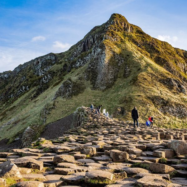 Visitors walking on the giant's causeway, with the characteristic hexagonal basalt columns and a rugged cliff in the background.