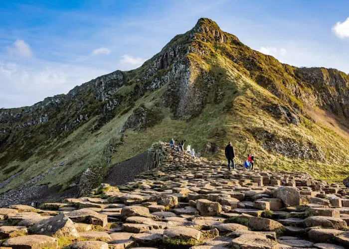 Guests on one of Overland Ireland’s small-group tours of Ireland walk on the breathtaking basalt hexagonal columns that make up the Giant’s Causeway in Northern Ireland. A green mountain rises to a sharp peak behind the natural wonder.