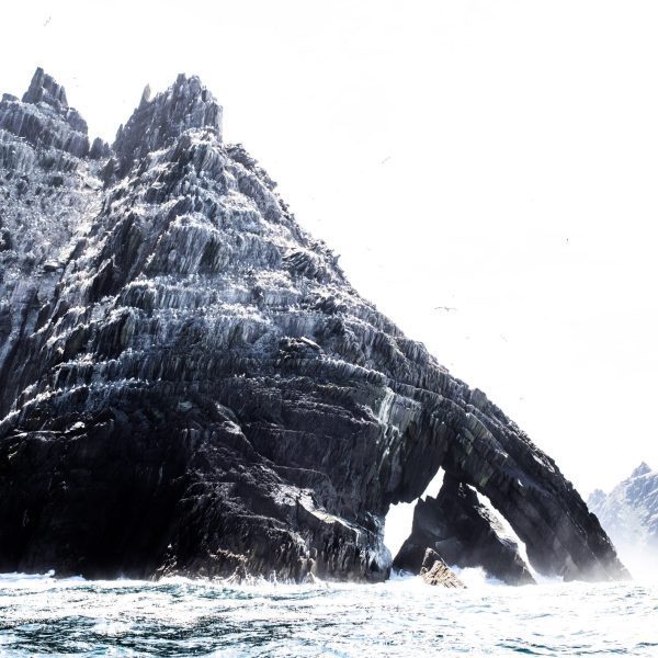 A rugged, snowy mountain emerges from the sea with a distinctive arch formation near its base.