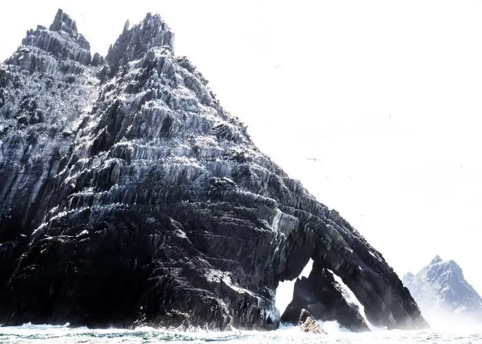 A rugged, snowy mountain emerges from the sea with a distinctive arch formation near its base.