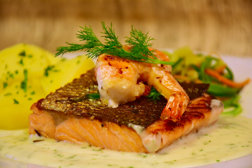 Grilled salmon fillet topped with a shrimp, served with a creamy sauce, fresh dill, and boiled potatoes on the side.