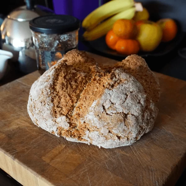 A freshly baked loaf of bread on a wooden cutting board with fruit and a teapot in the background.