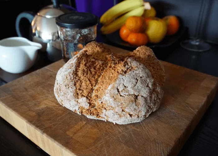 A freshly baked loaf of bread on a wooden cutting board with fruit and a teapot in the background.