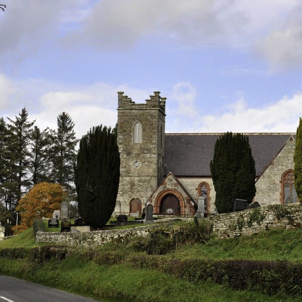 Historic stone church beside a road with a graveyard and trees under a cloudy sky.