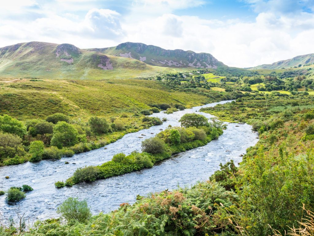 A serene river meandering through a lush, hilly landscape under a bright blue sky with clouds.