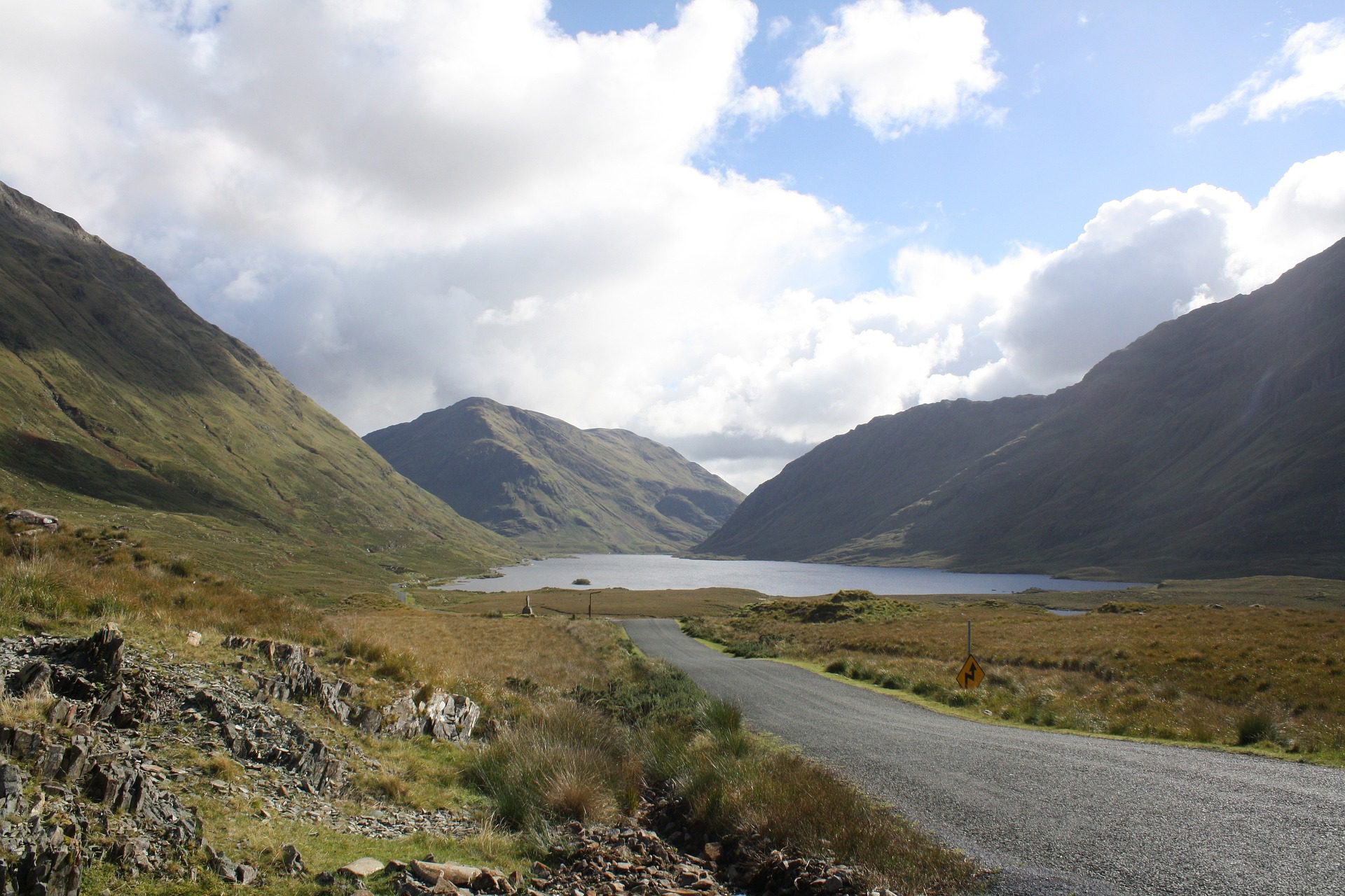 A scenic road winds through a valley with a lake nestled between rolling hills under a partly cloudy sky in Ireland.