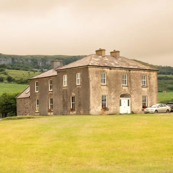 A two-story house with a weathered exterior, situated in a vast grassy field with hills in the background, under a cloudy sky.