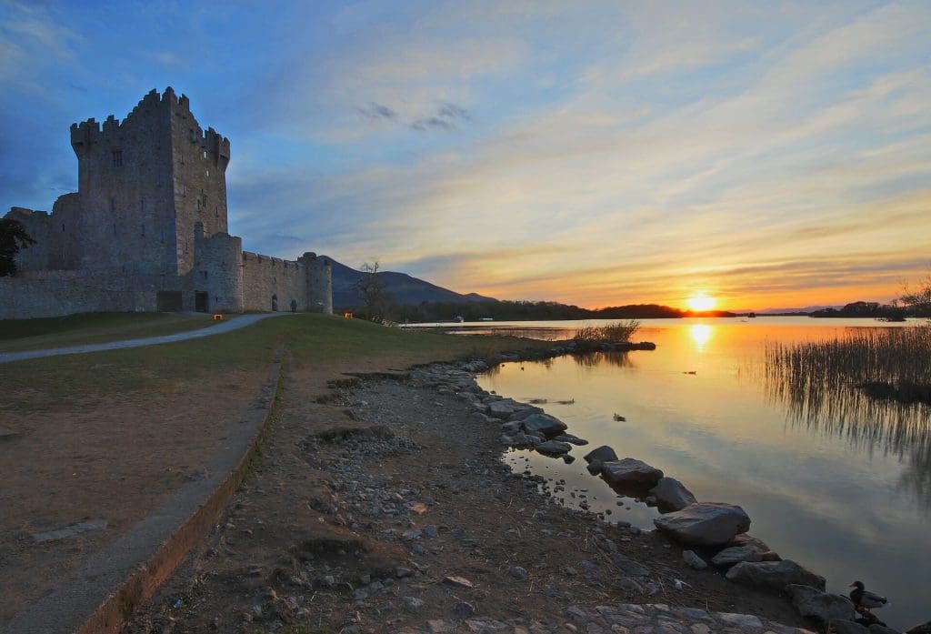 A medieval castle at sunset with calm waters in the foreground and a colorful sky in the background.