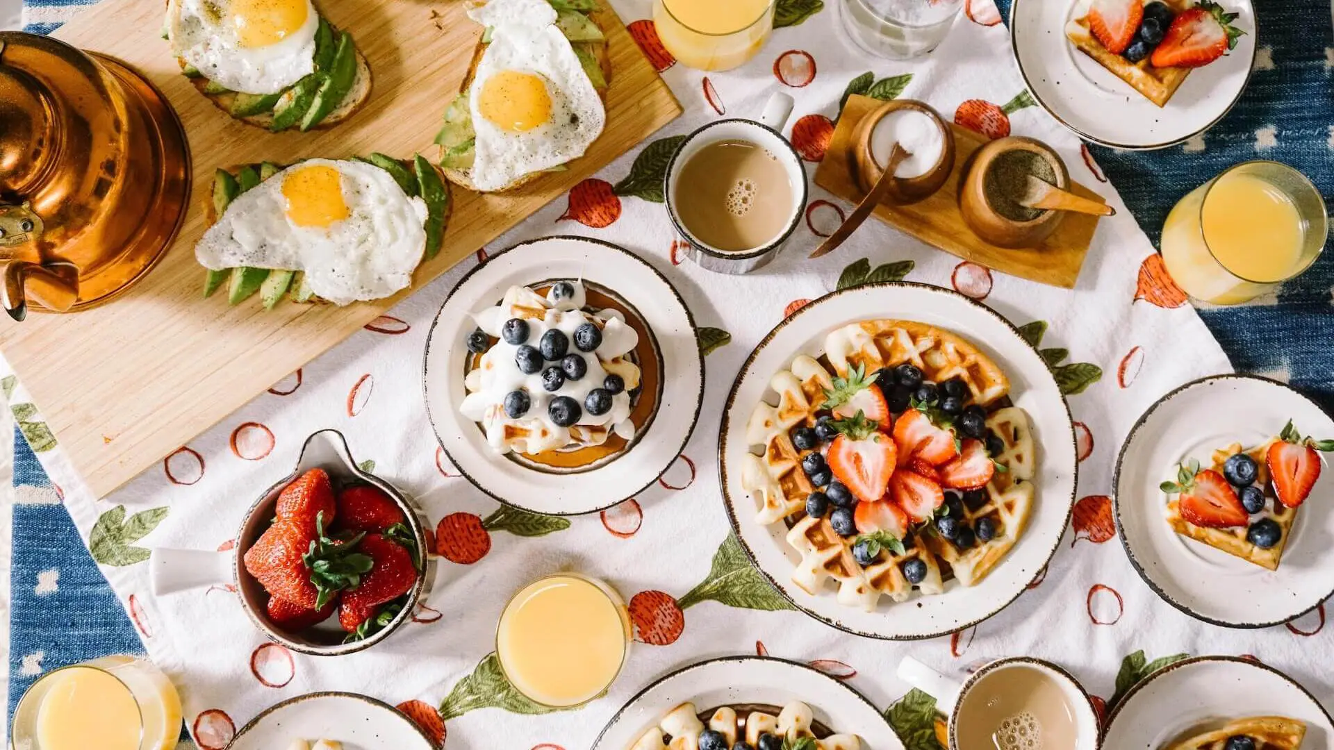 A breakfast spread featuring waffles with fresh berries, cups of coffee, and glasses of orange juice on a patterned tablecloth.