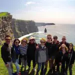 A group of guests on an Overland small-group Ireland tour pose for a photo on a sunny day with the famous Cliffs of Moher in County Clare, Ireland, behind them. The green grass covering the clifftops, the rugged cliff faces, and the roaring Atlantic Ocean in the background are quintessential images of Ireland’s Wild Atlantic Way.