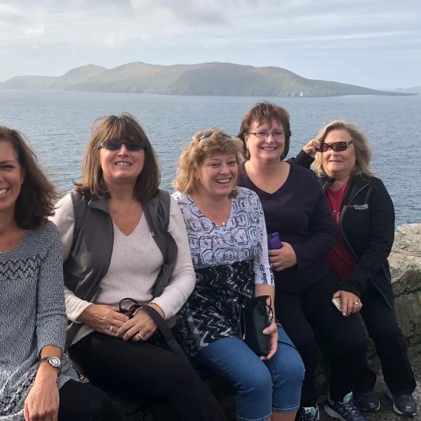 Five women sitting on a stone wall, smiling, with a scenic coastal view in Ireland behind them. A tranquil sea is on the other side of the wall, and out in the distance are rocky outcrops and islands. The ruggedly beautiful coastal view captures the essence of Overland’s small-group Ireland tours.