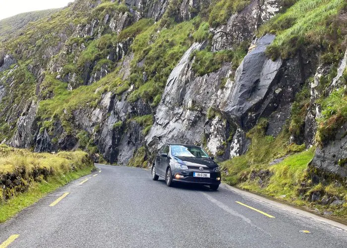A car parked on a narrow road with steep rocky cliffs on one side.