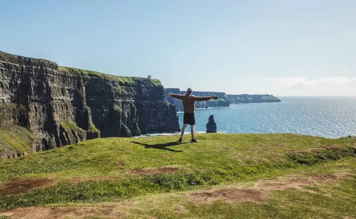 A person with outstretched arms standing on a grassy cliff overlooking the sea during a private tour.