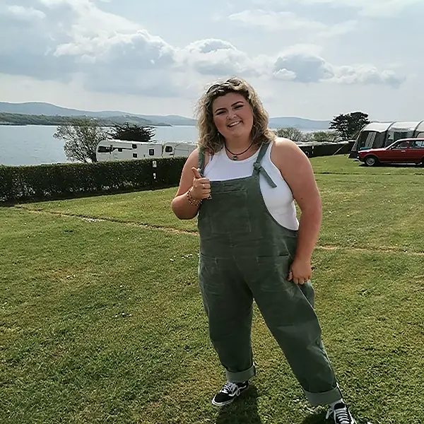 Cathy, standing on a grassy field with a thumbs-up gesture, a lake and caravans in the background on a sunny day.