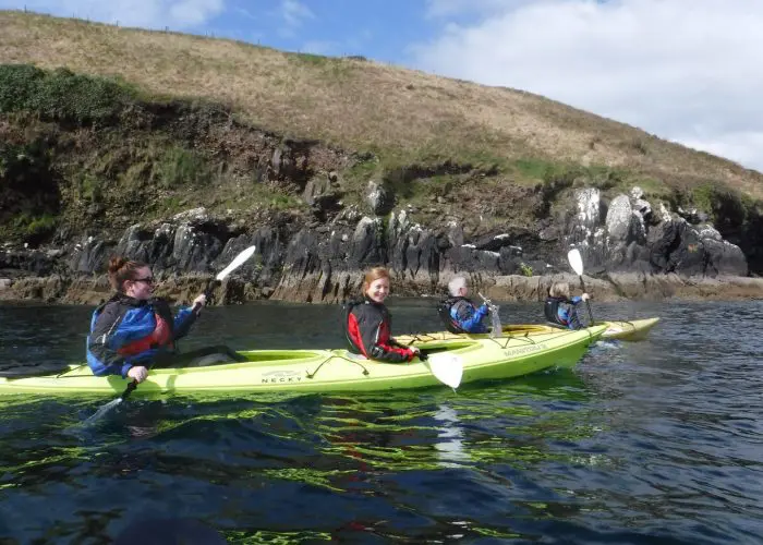 Three people in wetsuits kayaking near a rocky shore under a clear sky.