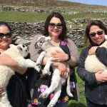 Three women smiling and holding lambs in a sunny field surrounded by stone walls.