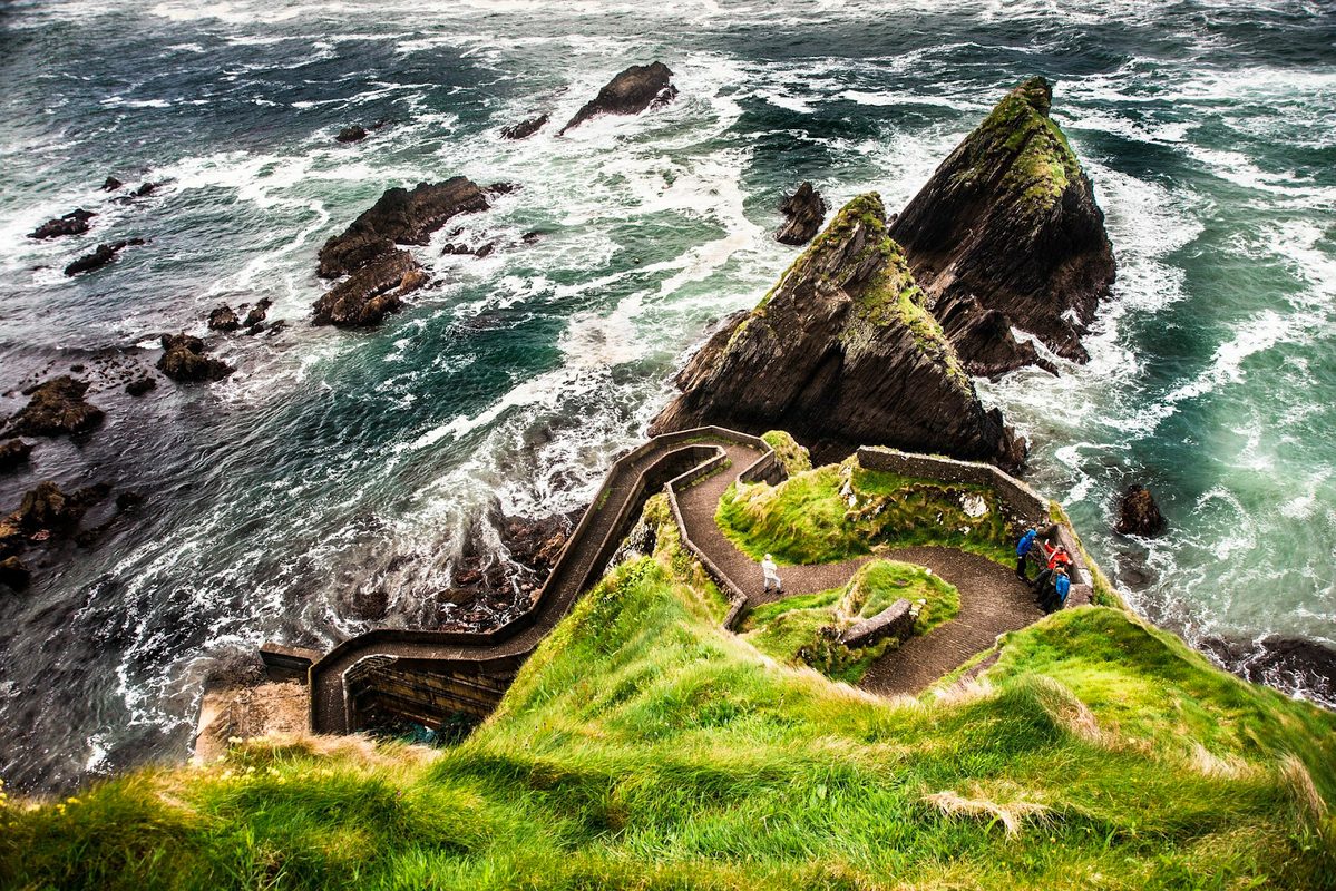 Coastal view of rugged cliffs with a winding stone path descending to the sea, two visitors visible amid vibrant green grass. waves crash against sharp rocks below.