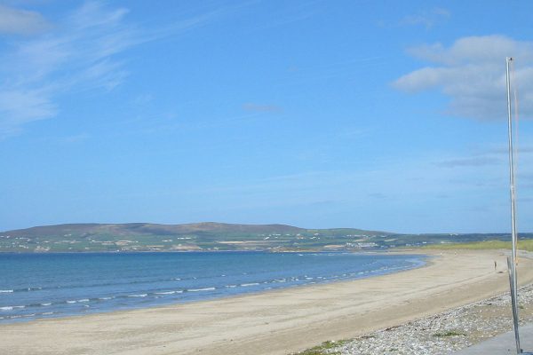 Empty beach with a curved shoreline, calm sea, and clear sky. a distant hill across the water and a series of poles on the left.