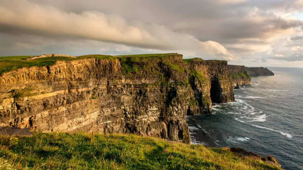 Lush green cliffs drop steeply to meet the ocean under a dramatic sky at sunset.