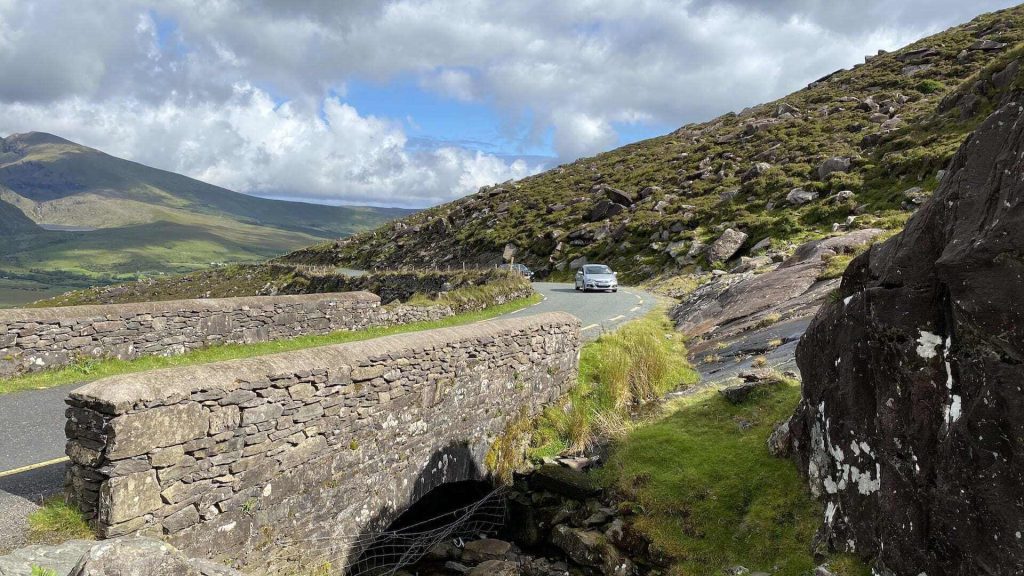 A car winding along narrow roads in a mountainous landscape and crossing a stone bridge under a partly cloudy sky.