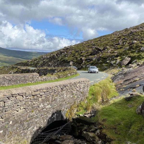 A car winding along narrow roads in a mountainous landscape and crossing a stone bridge under a partly cloudy sky.