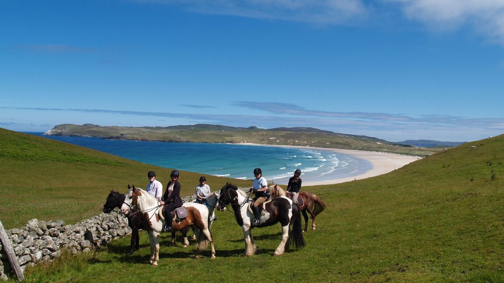 Group of riders on horses pausing near a beach with clear skies and a sweeping view of the ocean and hilly terrain.