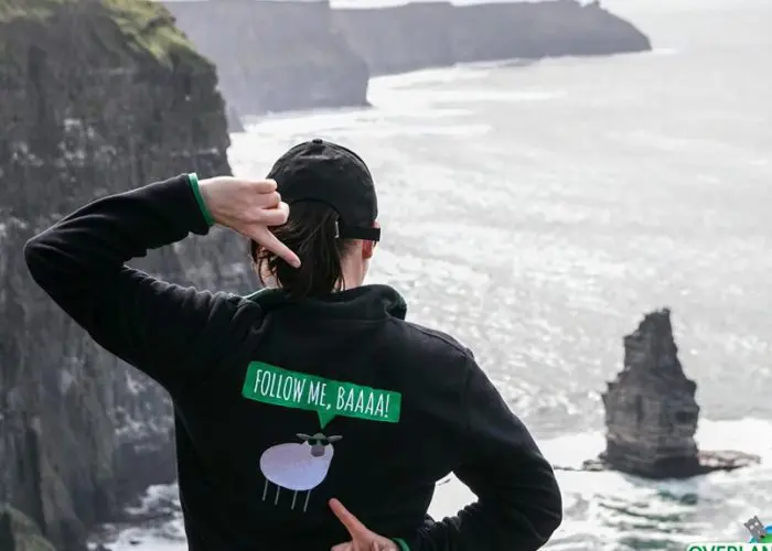 Person pointing at their hoodie, which features a cartoon sheep and the text "FOLLOW ME, BAAAA!" while standing on a cliffside overlooking the ocean.