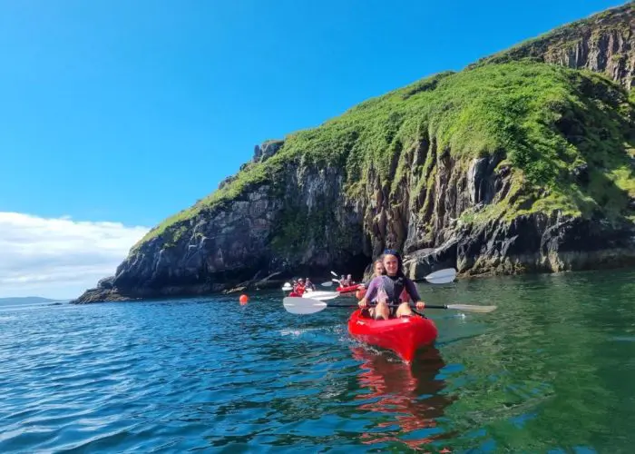 Two people kayaking near a rocky cliff covered with green vegetation under a clear blue sky.