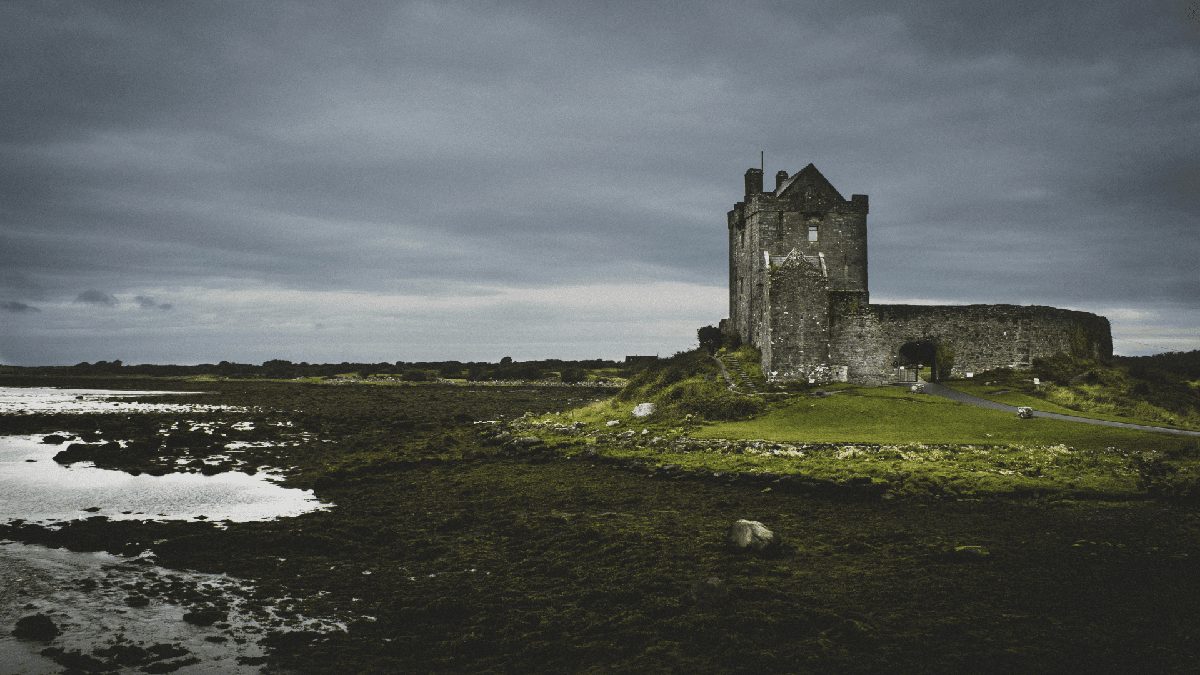 An ancient stone castle stands on a grassy coastline under a cloudy sky, with a calm sea visible to the left.