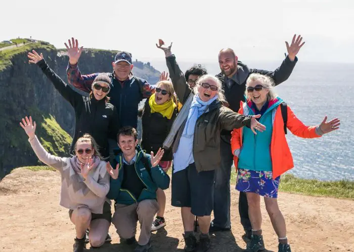 A group of joyful tourists with raised hands, posing for a photo at the cliffs of moher under a clear blue sky.