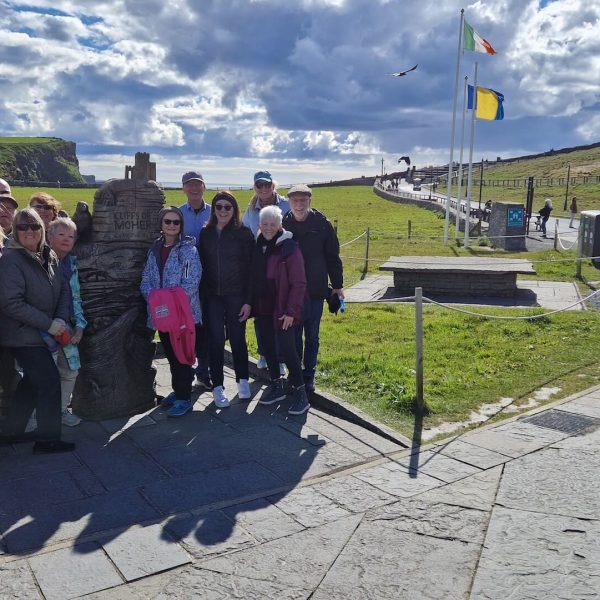 A group of people stands next to a stone monument on a paved path with flags in the background, under a partly cloudy sky in an open landscape, capturing the essence of an Ireland tour package.