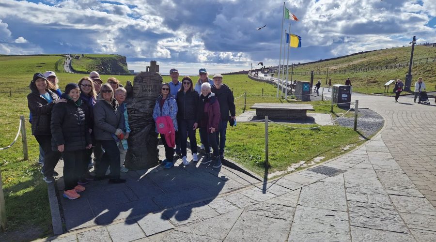 A group of people stands next to a stone monument on a paved path with flags in the background, under a partly cloudy sky in an open landscape, capturing the essence of an Ireland tour package.