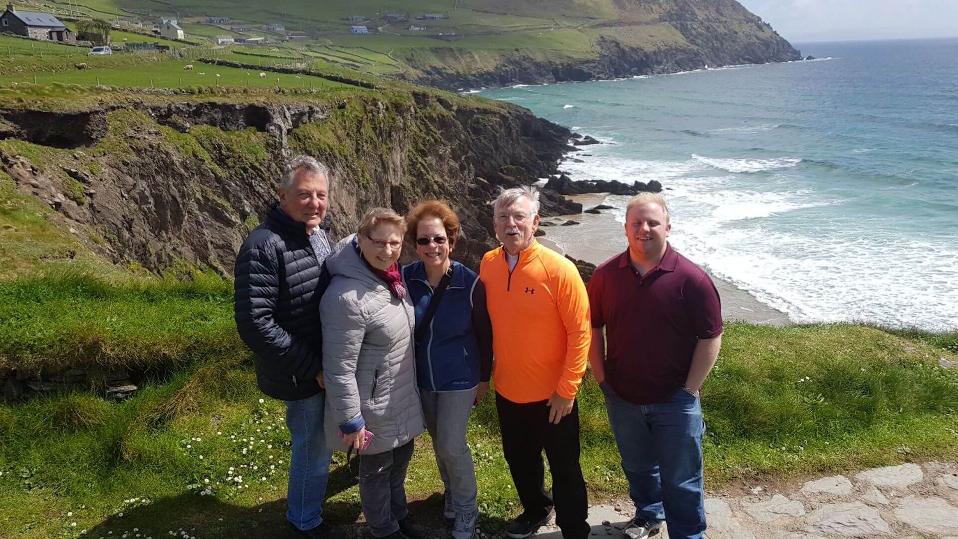 A group of five people stand on a grassy cliff overlooking a rocky coastline and ocean, with green hills in the background, capturing the essence of an unforgettable Ireland itinerary 7 days.