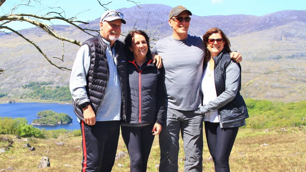 Four people are standing together outdoors, dressed casually, with a scenic background of Ireland's mountains, greenery, and a lake. They appear happy and are posing for the camera. Looks like they just finished an Active Tour!